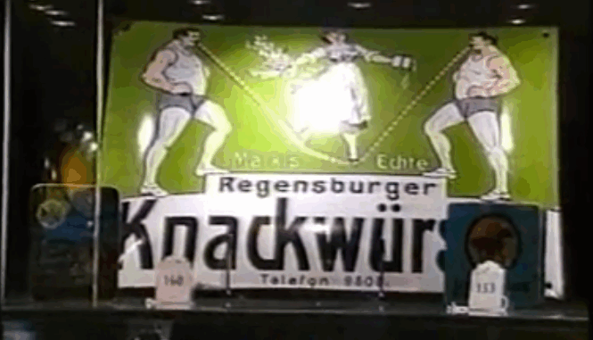 Ein altes grün-weißes Schild mit der Aufschrift "Regensburger Knackwürstchen" ist hinter Glas als Teil einer Sammlung ausgestellt. Zwei muskulöse Männer in Ringerhosen stehen sich gegenüber, zwischen ihnen ein Blumenstrauß, flankiert von kleinen Plaketten mit Nummern.