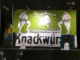 Ein altes grün-weißes Schild mit der Aufschrift "Regensburger Knackwürstchen" ist hinter Glas als Teil einer Sammlung ausgestellt. Zwei muskulöse Männer in Ringerhosen stehen sich gegenüber, zwischen ihnen ein Blumenstrauß, flankiert von kleinen Plaketten mit Nummern.