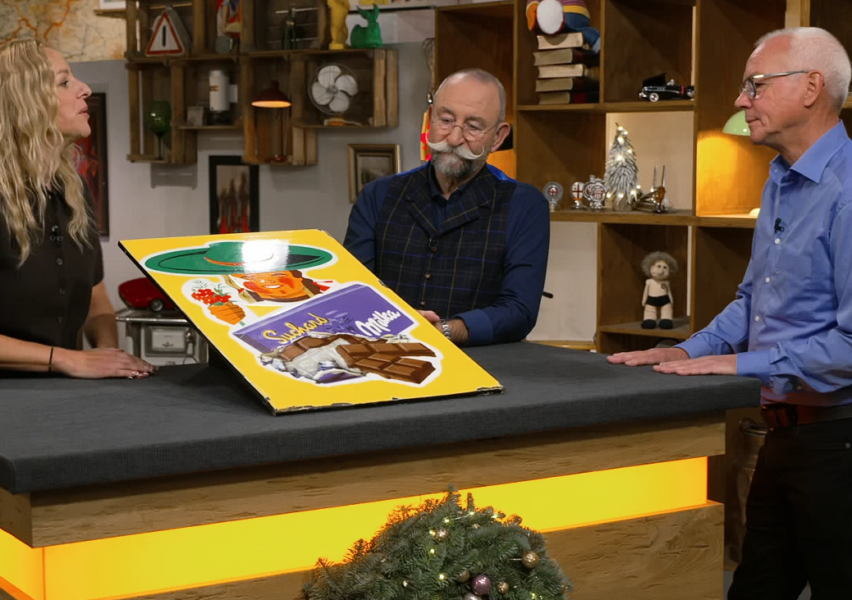 Zu dieser Illustration: Drei Erwachsene betrachten eine große, alte Milka-Schokoladenwerbung auf einem Tisch, die die schokoladige Erfolgsgeschichte von Philippe Suchard inmitten von Atelierregalen und verschiedenen Dekorationen im Hintergrund feiert. (Alt-Text realisiert durch KI)