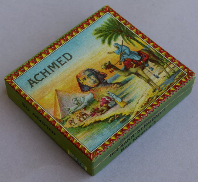 Eine dekorative Schachtel mit aegyptischem Motiv aus dem 20. Jahrhundert Der Deckel zeigt eine lebendige Wuestenszene mit Pyramiden, einem Kamel und Palmen. ACHMED ist deutlich sichtbar eingerahmt von einem gruenen Rand und einem rot-gelben Muster an den Raendern ein Schmuckstueck aus Bad Kreuznach.