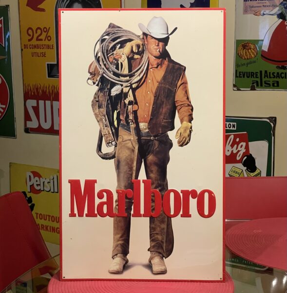 Ein altes Marlboro-Werbeschild mit dem kultigen Marlboro-Man-Cowboy mit weißem Hut und brauner Weste, der ein aufgerolltes Seil und eine Ausrüstung in der Hand hält, mit "Marlboro" in fetten roten Buchstaben am unteren Rand. Retro-Poster tragen zum nostalgischen Charme dieses Kultobjekts bei.