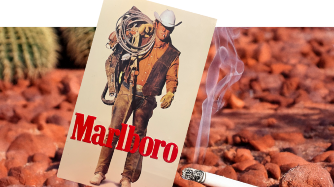 Eine gedruckte Marlboro-Werbung mit dem ikonischen Marlboro-Man-Cowboy, der ein Lasso hält, ist zwischen rötlich-braunen Felsen aufgehängt. Davor raucht eine angezündete Zigarette, während Kakteen den Hintergrund bevölkern.