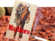 Eine gedruckte Marlboro-Werbung mit dem ikonischen Marlboro-Man-Cowboy, der ein Lasso hält, ist zwischen rötlich-braunen Felsen aufgehängt. Davor raucht eine angezündete Zigarette, während Kakteen den Hintergrund bevölkern.