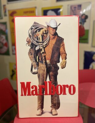 Ein Mann mit Cowboyhut, Weste und Handschuhen trägt einen Sattel und ein Seil über seiner Schulter. Das Wort "Marlboro" steht in großen roten Buchstaben am unteren Rand. Im Hintergrund dieser klassischen Kleinanzeige sind alte Plakate zu sehen.