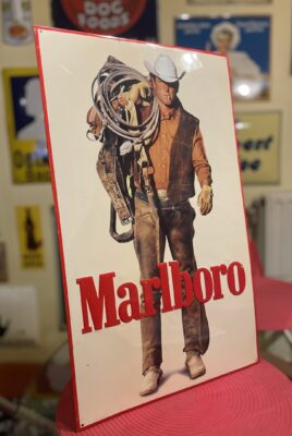 Eine alte Marlboro-Werbung, die einen Cowboy mit Hut und Westernkleidung zeigt, der ein Seil und Handschuhe in der Hand hält, mit einem fetten roten "Marlboro"-Text am unteren Rand. Dieses ikonische, klassifizierte Plakat erscheint zusammen mit anderen im unscharfen Hintergrund.