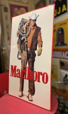 Ein Metallschild mit einem Cowboy mit weißem Hut und brauner Weste, der ein aufgerolltes Seil und einen Sattel hält, mit dem Wort "Marlboro" in großen roten Buchstaben am unteren Rand. Dieser klassifizierte Artikel ist auf einem roten Stuhl in einem Raum ausgestellt.