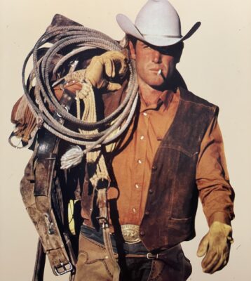 Ein Mann mit einem weißen Cowboyhut, einer braunen Weste, einem orangefarbenen Hemd und gelben Handschuhen trägt ein aufgerolltes Seil und einen Sattel über der Schulter. Mit einer Zigarette im Mund blickt er nach vorne, wie eine klassifizierte Figur aus einer Auto Draft-Auswahl.