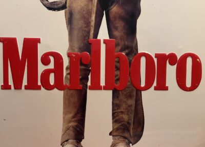 Roter Marlboro-Text mit der Aufschrift "Classified" überlagert das Bild einer Person in braunen Lederchaps und Stiefeln; nur der Unterkörper ist sichtbar, wobei der Text die Chaps teilweise verdeckt.