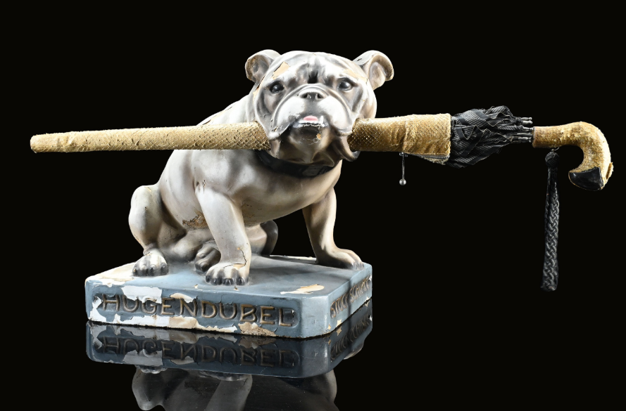 Hugendubel-Werbefigur „Bulldogge“ aus Gips, Deutschland, um 1930, Höhe 31 cm. Mit originalem Regenschirm; durch die Öffnungen in den Backen noch vollständig bestückt. Schönes Originalexemplar mit altersbedingten Lackabplatzungen. (Bild: Wormser Classic Auctions)